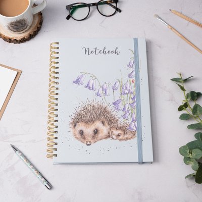 Two hedgehogs cuddling next to flowers on an A4 spiral bound notebook on a desk surrounded by stationery